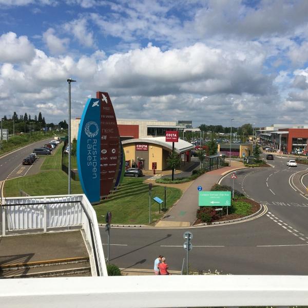Large sign for Rushden Lakes retail park with the road entering the site in the background.
